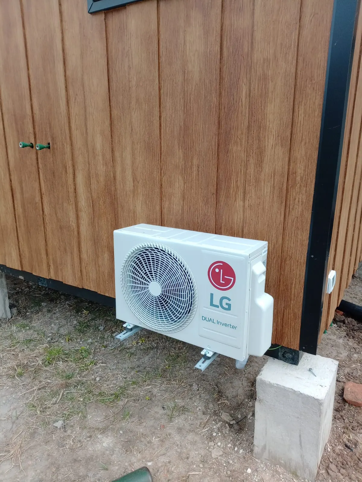 Unidad exterior LG Dual Inverter sobre soportes junto a una casa de madera